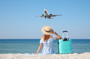 Woman,With,Suitcase,On,Sandy,Beach,Looking,At,Airplane,Flying